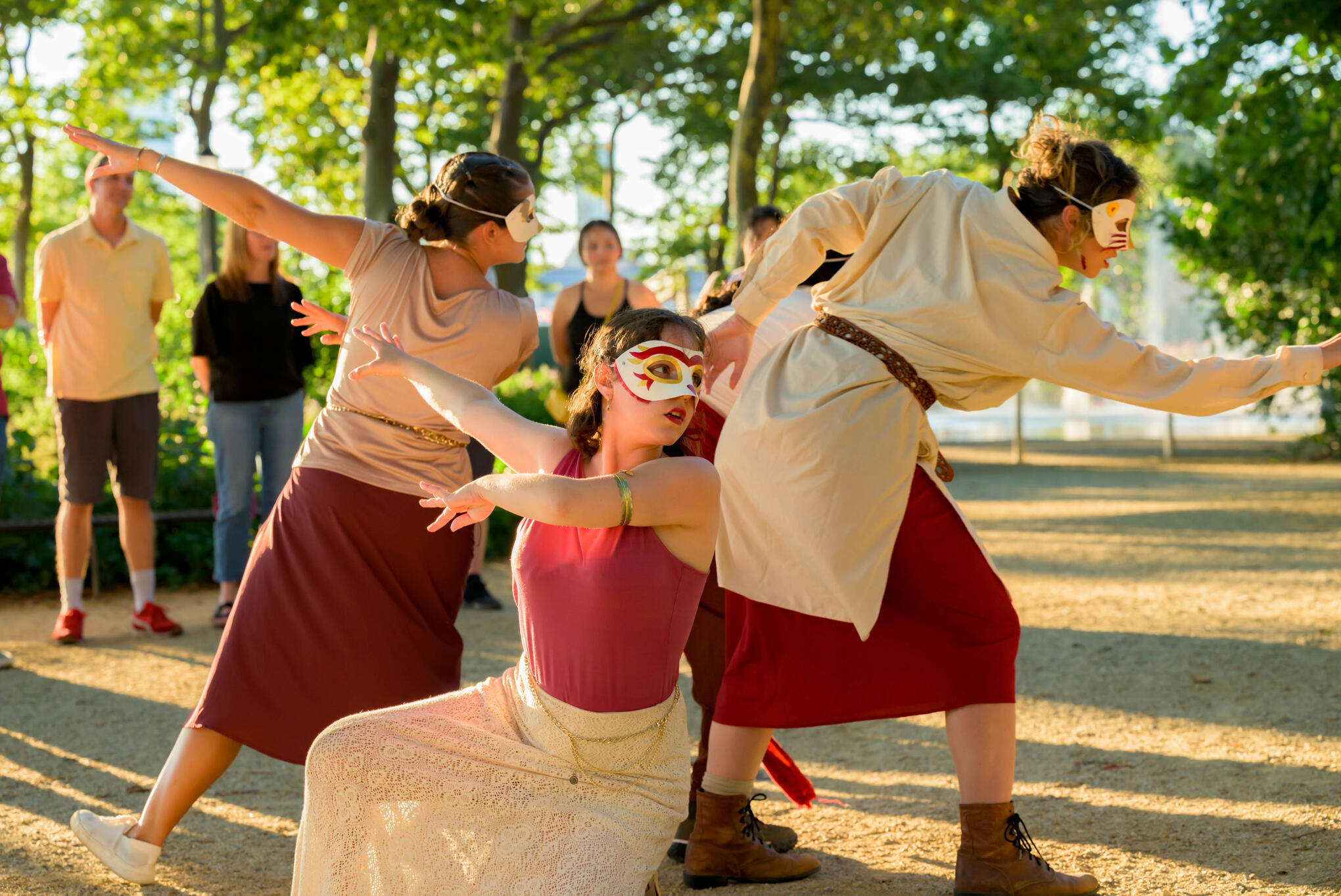 Sivan, a young female, is smiling and looking into the camera. She is standing in a park full of greenery that provides a blurry background. They are wearing a white tank top with a lacy design, and have brown wavy hair that is pulled back.