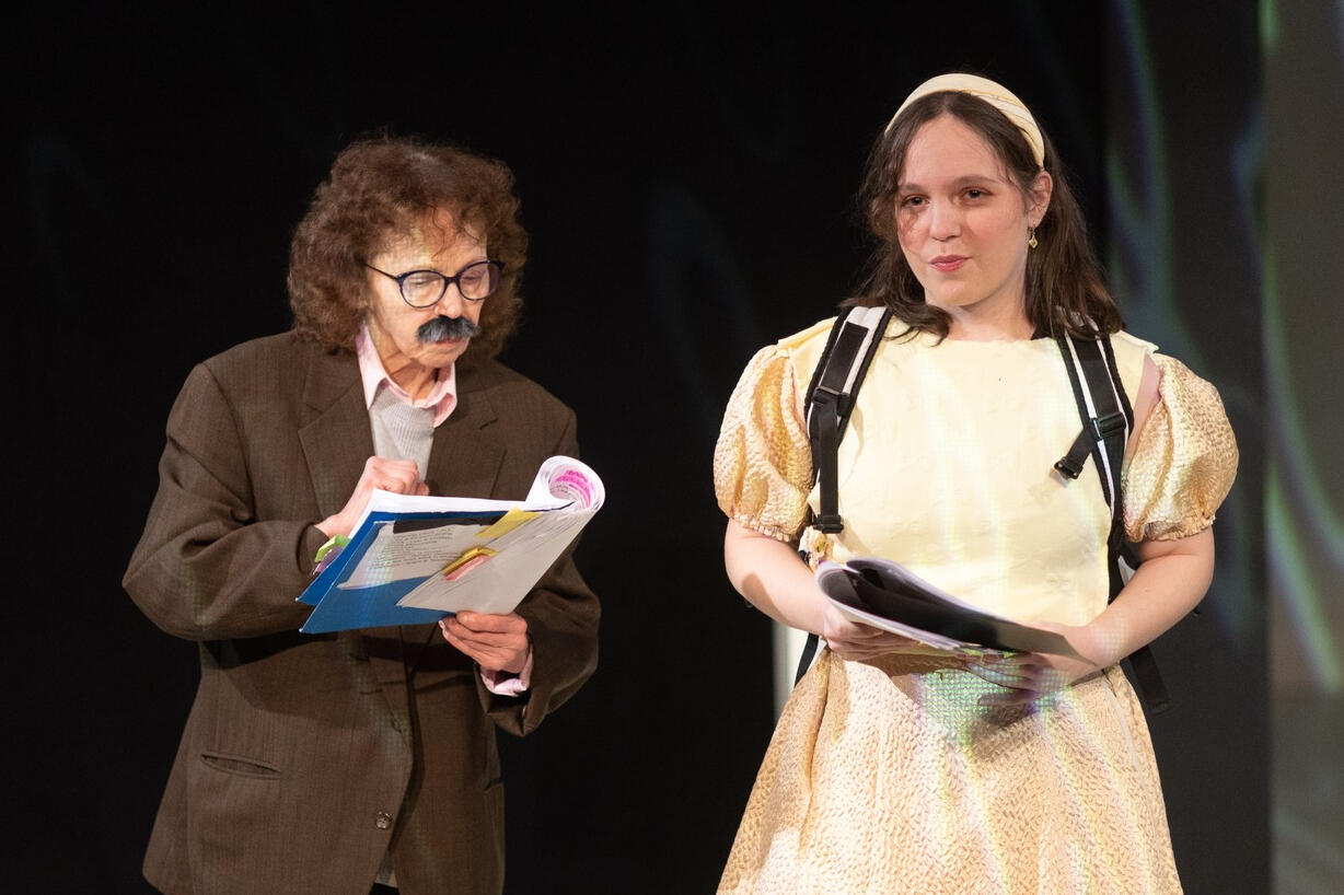 Sivan, a young female, is smiling and looking into the camera. She is standing in a park full of greenery that provides a blurry background. They are wearing a white tank top with a lacy design, and have brown wavy hair that is pulled back.