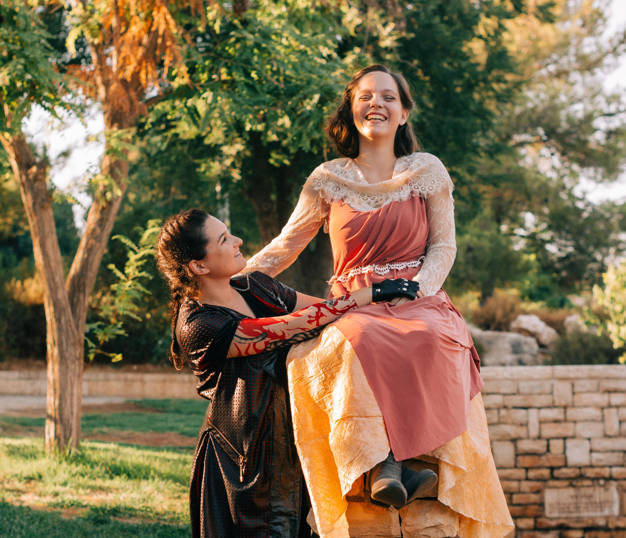 Sivan, a young female, is smiling and looking into the camera. She is standing in a park full of greenery that provides a blurry background. They are wearing a white tank top with a lacy design, and have brown wavy hair that is pulled back.