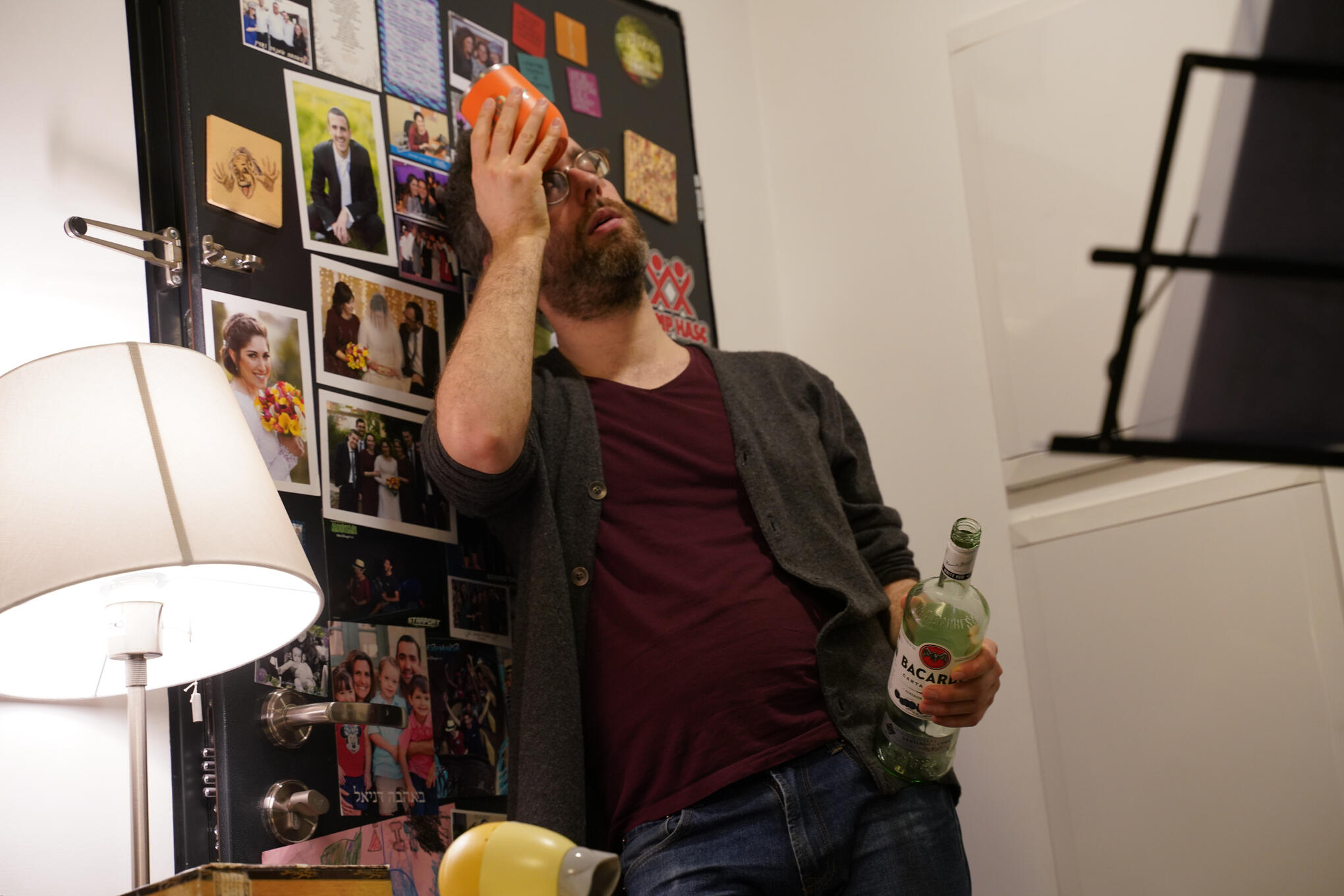 Sivan, a young female, is smiling and looking into the camera. She is standing in a park full of greenery that provides a blurry background. They are wearing a white tank top with a lacy design, and have brown wavy hair that is pulled back.